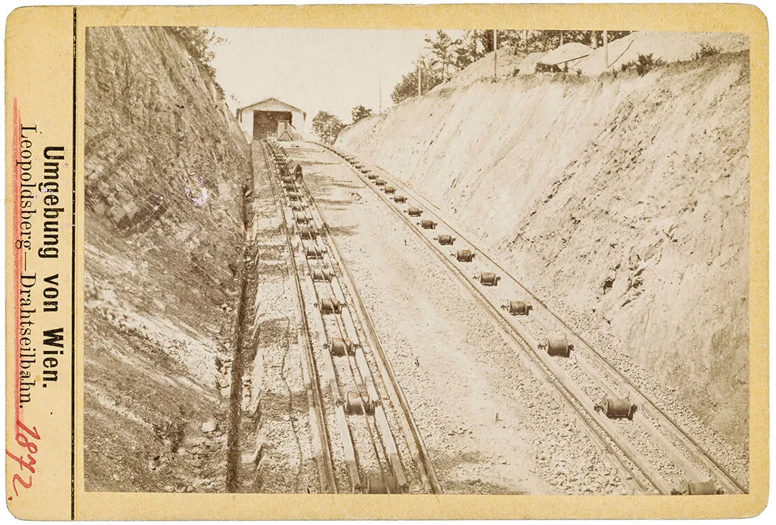 Drahtseilbahn auf den Kahlenberg, Blick auf die Trasse Richtung Bergstation, Fotografie, vermutlich von Michael Frankenstein, um 1873/75, Wien Museum. „Der gute Kahlenberg wird nun auch schon von der weiterschreitenden Kultur beleckt. Ein Schienenstrang zieht sich bereits über den Rücken des Berges hinauf und schwere Waggons mit lustigen Wienern und Wienerinnen angepfropft, wird er von morgen an zu tragen bekommen. Der arme Kahlenberg! hat sich's gewiß nicht träumen lassen, daß er im Jahre 1873 „Aktiengesellschafts-Bildungsobjekt" werden wird!“ (Illustrirtes Wiener Extrablatt, 27. Juli 1873, S. 2)