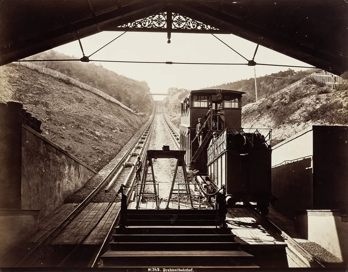 Drahtseilbahn auf den Kahlenberg, Blick aus der Talstation auf die Trasse, Fotografie von Michael Frankenstein, um 1873/75, Wien Museum. Die Aufnahme zeigt den Blick aus der Talstation auf die Trasse mit der diese im oberen Streckenabschnitt überspannenden Bründlwegbrücke. An den Personenwagon angehängt ist der Tenderwagen, der benutzt wurde, um Wasser und Kohle für das Kesselhaus zu transportieren. Die Beladung des Tenderwagens erfolgte über die hier rechts und links zu sehenden Rampen.