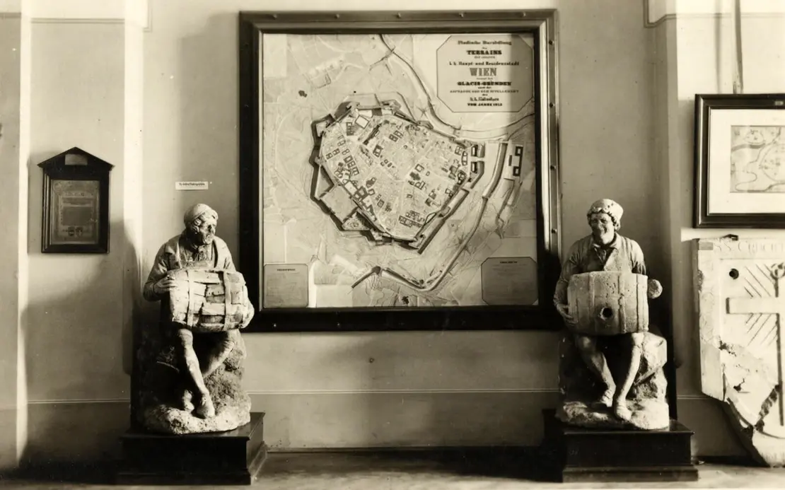 Ausstellungsraum des Historischen Museums der Stadt Wien im Rathaus, Postkarte, um 1930, Wien Museum. Reliefplan von Wien und Hauszeichen „Zum schönen Binder" vom Haus Siebensterngasse 34 (abgebrochen 1867, um 1770). Die beiden Skulpturen aus Sandstein – besonders aufwendige Hauszeichen – wurden 1914 vom Historischen Museum erworben.